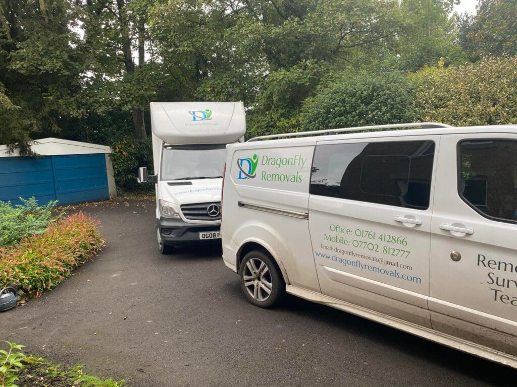 Two white removal vans from "DragonFly Removals" are parked on a driveway surrounded by trees and greenery. One van displays contact information, and a garage with a blue door is visible in the background.