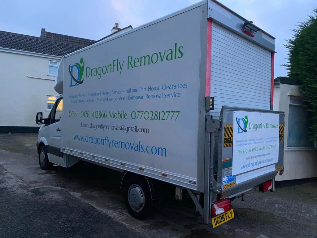 A white removal van with "DragonFly Removals" branding is parked in a driveway. The van's side displays contact details, including phone numbers and email, along with a logo of a dragonfly.