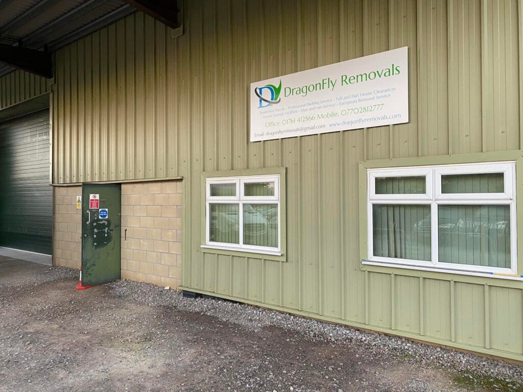 Exterior of a beige warehouse building with a sign reading "Dragonfly Removals." The building has a green garage door, two white-framed windows, and a small entrance door on a gravel surface.