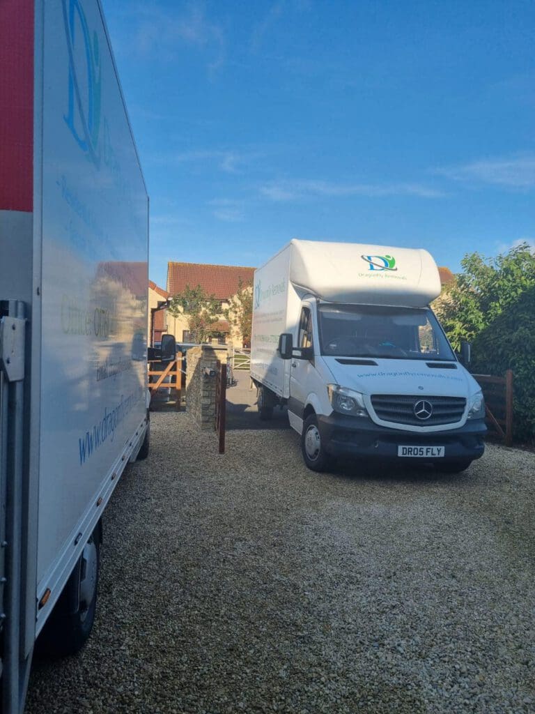 Two white moving trucks are parked in a driveway. The truck on the right is facing forward, while the one on the left is partially visible. The background shows a blue sky, trees, and part of a house with a red roof.