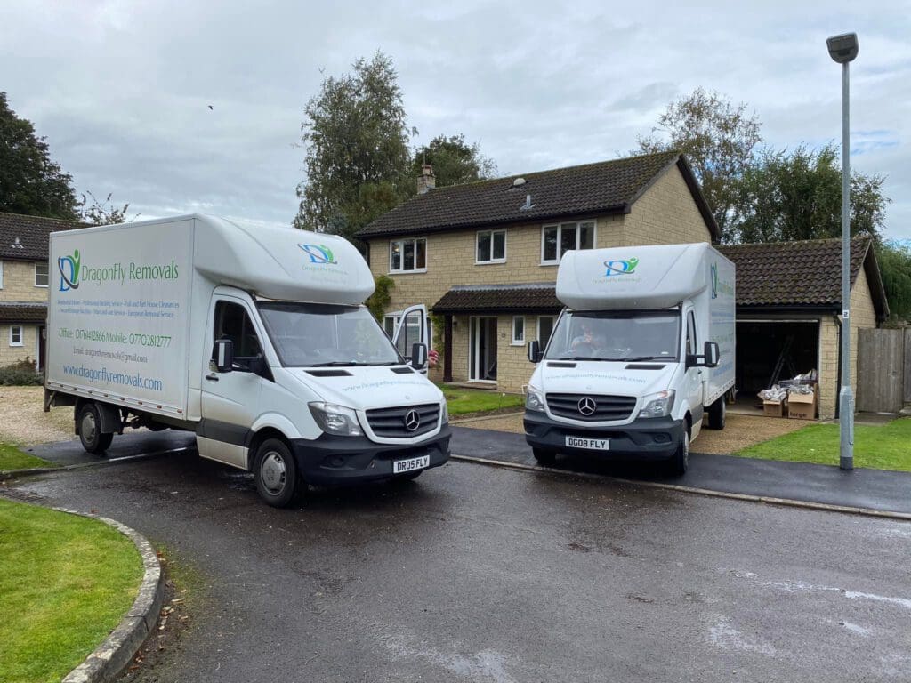 Two white moving vans with "Dragonfly Removals" branding are parked on a residential street in front of a beige two-story house. The vans have Mercedes-Benz logos. The weather is cloudy, and trees are visible in the background.