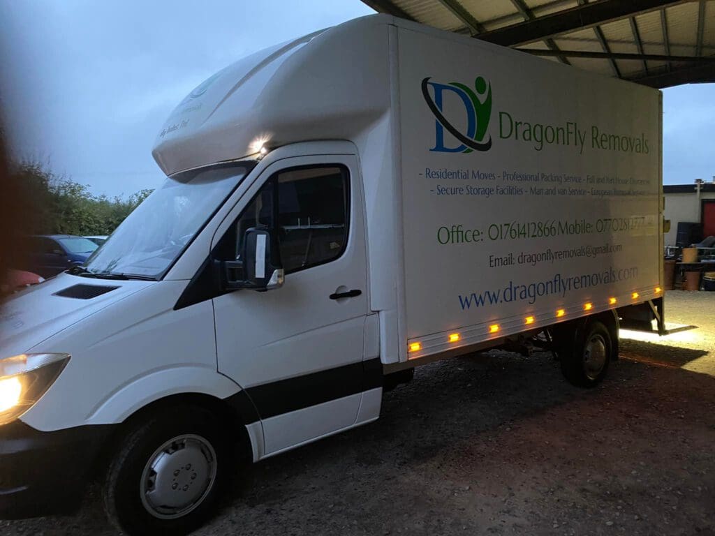 A white moving truck with "DragonFly Removals" and services like "Residential Moves" printed on the side. The vehicle is parked under a shelter, and the headlights are on. Contact information and the website are also visible on the truck.