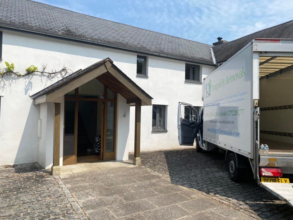 A moving truck is parked outside a white house with an open door. The truck is labeled "Dragonfly Removals." The house has a tiled roof and a wooden porch. It's a sunny day with a blue sky visible.