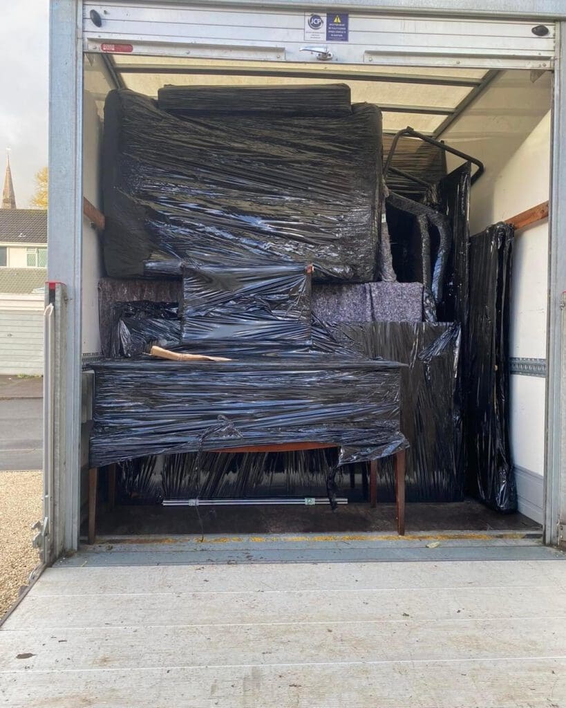 A moving truck loaded with furniture wrapped in black plastic for protection. The items are tightly packed and stacked inside the vehicle, including a large table with a wooden handle poking out in the center. The truck is parked outdoors.