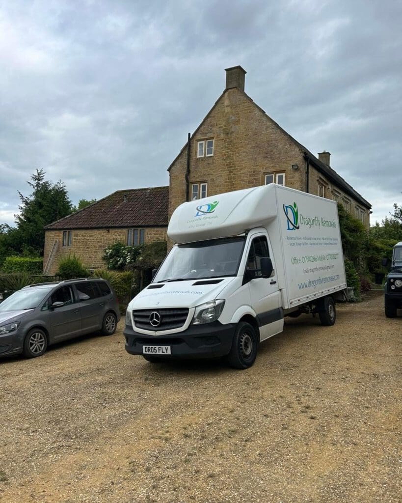 A white Mercedes moving van with "Dragonfly Removals" branding is parked on a gravel driveway in front of a large stone house. Other vehicles are parked nearby. The sky is overcast.