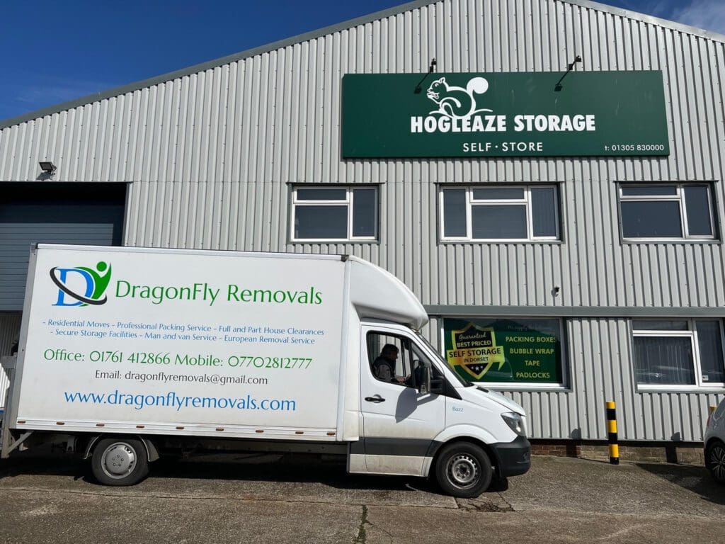 A white moving truck with "DragonFly Removals" branding is parked in front of a corrugated metal building labeled "Hogleaze Storage Self-Store." The truck displays contact information for the removals company.