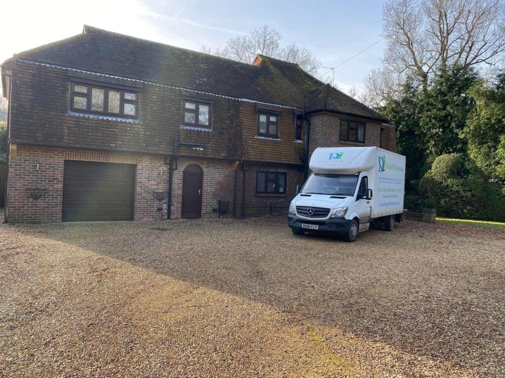 A large brick house with a brown tiled roof and a garage is pictured. A white delivery truck is parked in the driveway. The truck has green and blue writing on the side. There are trees and bushes around the house.