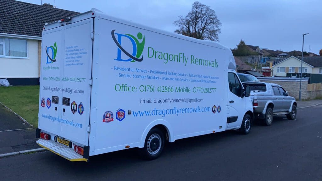 A white removal van with "DragonFly Removals" branding and contact details parked on a residential street. The van advertises services such as residential moves and secure storage. A white pickup truck is parked in front of the van.