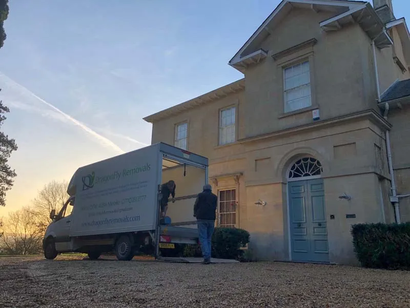 A person stands near the back of a removal van, which is parked in front of a large, beige house with an arched doorway. The van has "Proactive Removals" written on the side. The sun is setting, casting a warm glow over the scene.