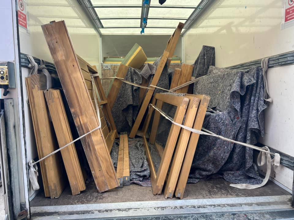 Wooden frames are secured with straps inside a truck. The frames are surrounded by protective padding, creating an organized yet cluttered appearance within the vehicle. The truck's interior walls and ceiling are visible.