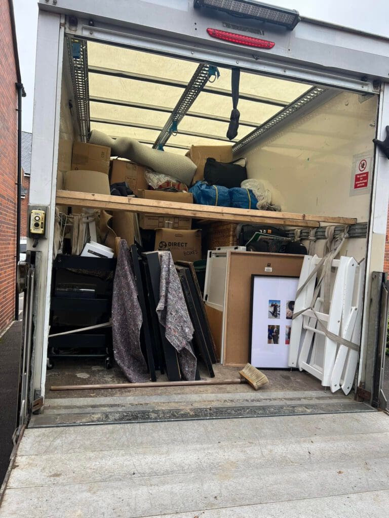 Open back of a moving truck filled with various packed items, including boxes, rolled carpets, picture frames, and other household goods. Some items are stacked on a wooden shelf and the truck is parked near a brick building.