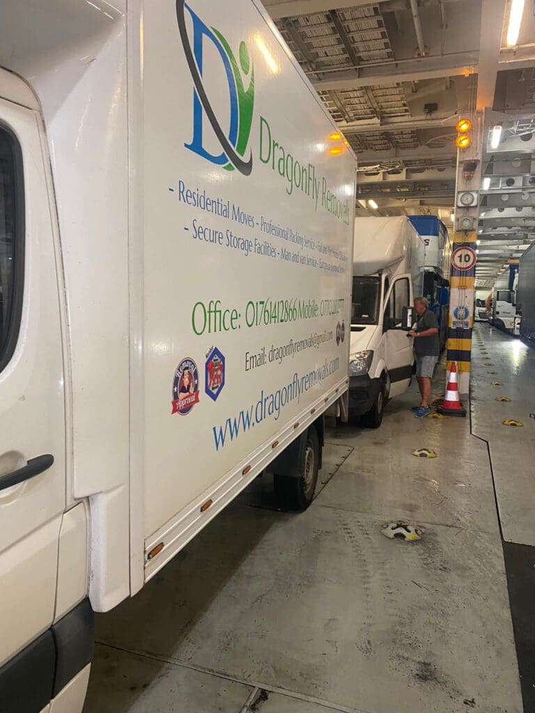 A moving van with "DragonFly Removals" logo and contact information is parked inside a large ferry or cargo ship with other vehicles. A person in casual attire stands nearby, looking at one of the vans. Various safety signs and equipment are visible.