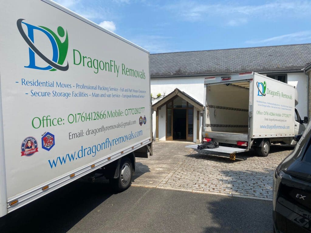 Two DragonFly Removals trucks parked outside a building, with one truck open. The company specializes in residential moves and packing services. Contact details are visible on the truck. The sky is clear and sunny.