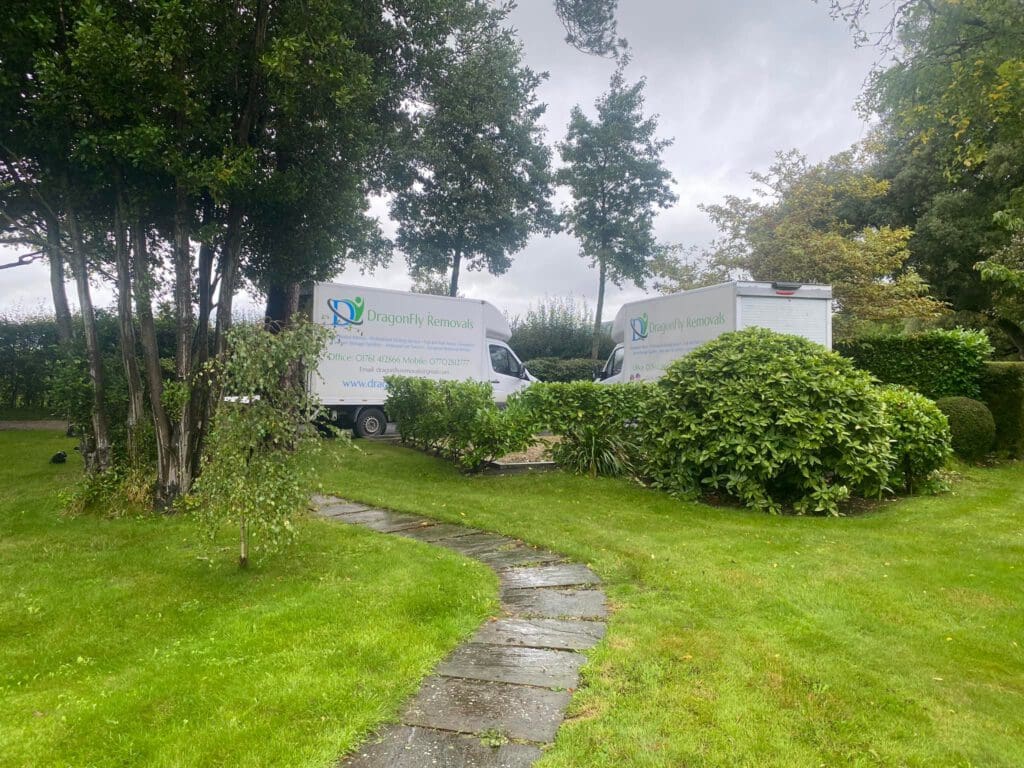 Two white moving vans with "Dragonfly Removals" logos are parked on a grass lawn surrounded by lush greenery and trees. A stone path leads through the garden, and the sky is overcast.