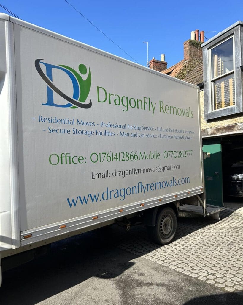 A white moving truck displays a logo and the name "DragonFly Removals." It lists services like residential moves and packing. Contact information includes a phone number and email. The truck is parked on a cobblestone street next to buildings.