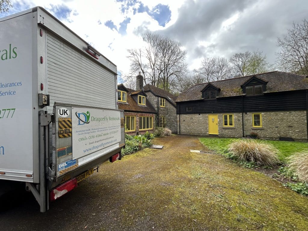 A moving truck with "Dragonfly Removals" on the side is parked on a driveway in front of a stone house with black and yellow accents. The surrounding area has grass and shrubs, and trees are visible in the background under a cloudy sky.