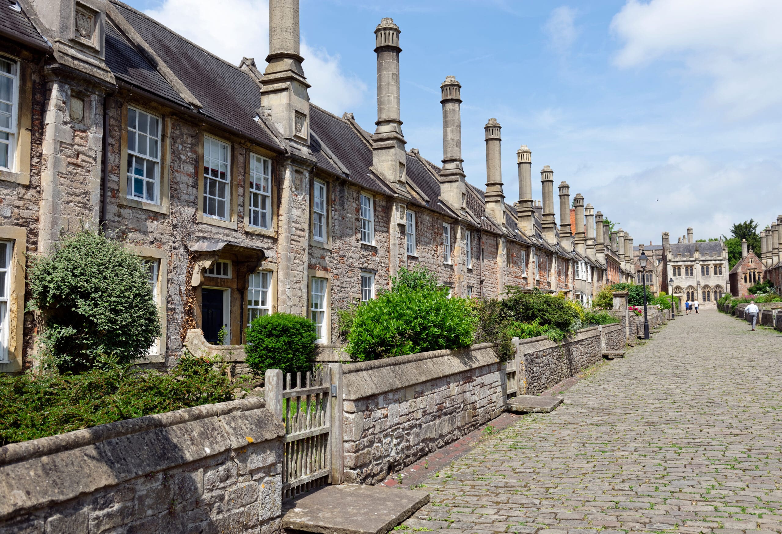 A row of historical stone cottages with tall chimneys and lush greenery at the front. The cobblestone street leads towards a distant building under a blue sky with scattered clouds.