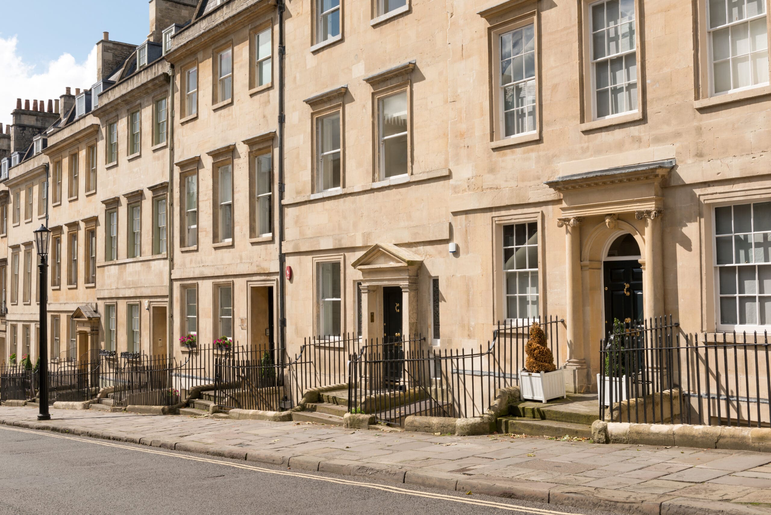 A row of Georgian-style terraced houses with light-colored stone facades, tall windows, and black iron railings. The road in front is empty, with stone-paved sidewalks and clear skies above.