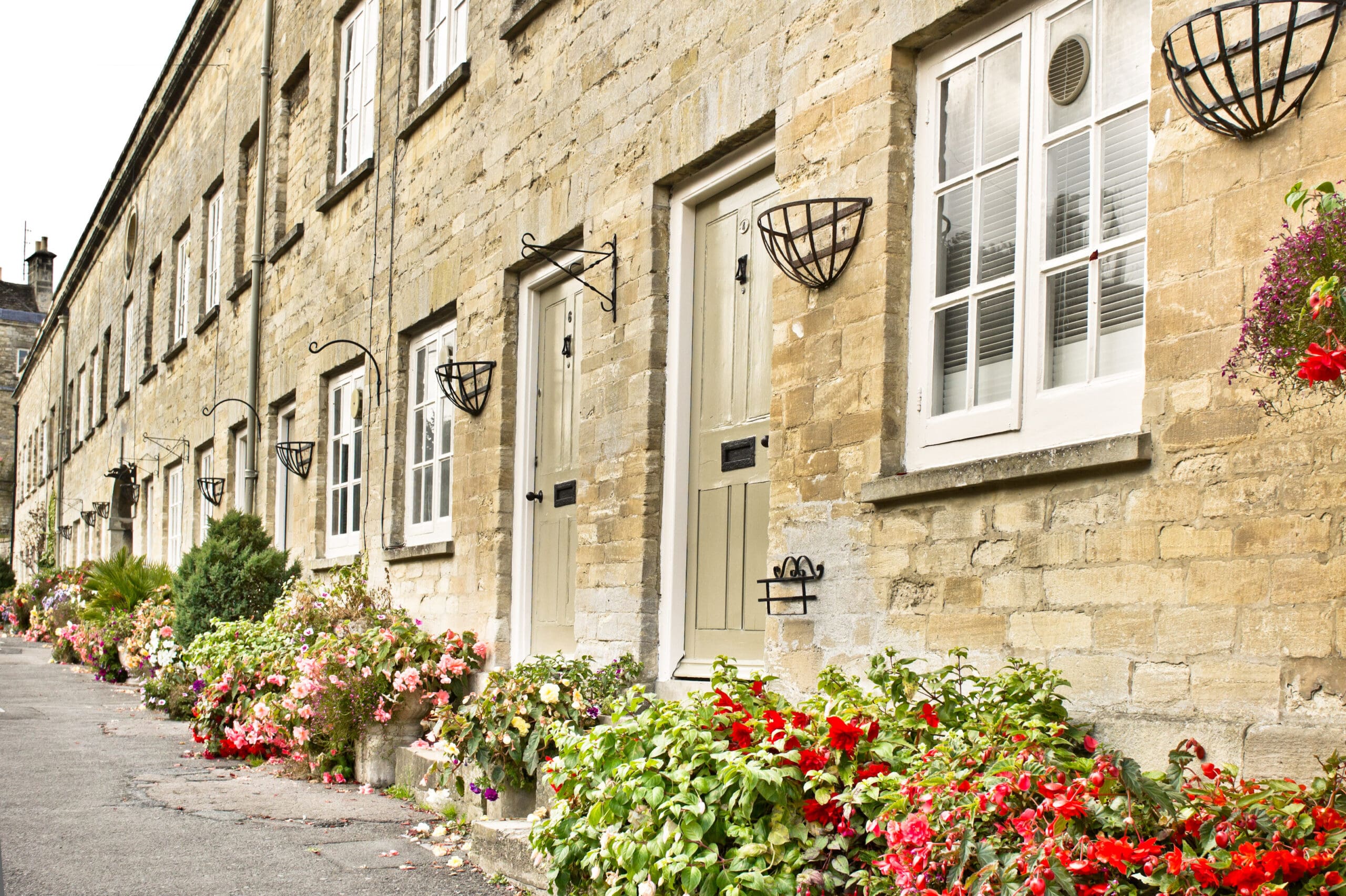 A row of charming stone terrace houses with white-framed windows and doors. Colorful flowers in full bloom adorn the fronts, creating a vibrant and welcoming atmosphere along the narrow street.