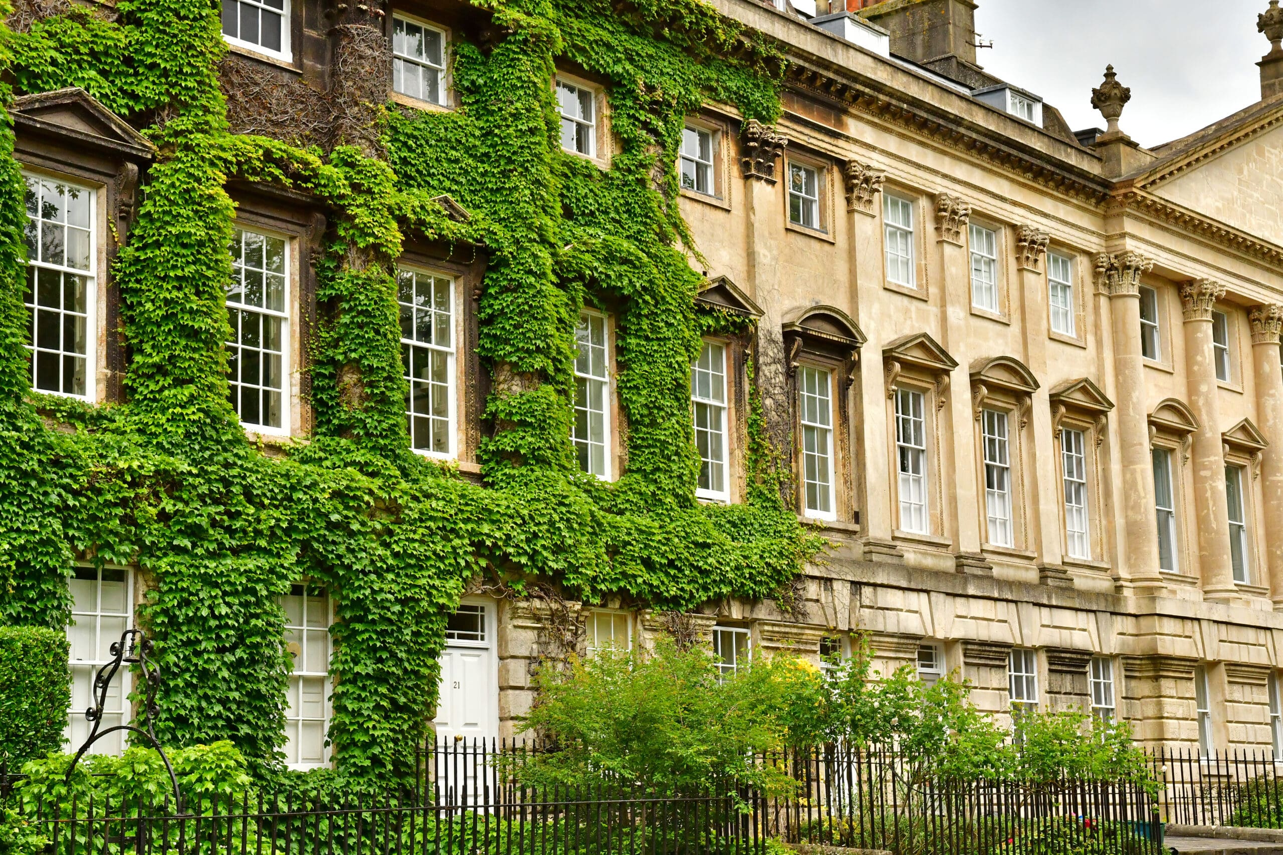 Historic row of stone buildings with tall windows. The left building is covered in lush green ivy, contrasting with the bare stone of the adjacent structure. A black wrought-iron fence and shrubbery line the front. Cloudy sky overhead.