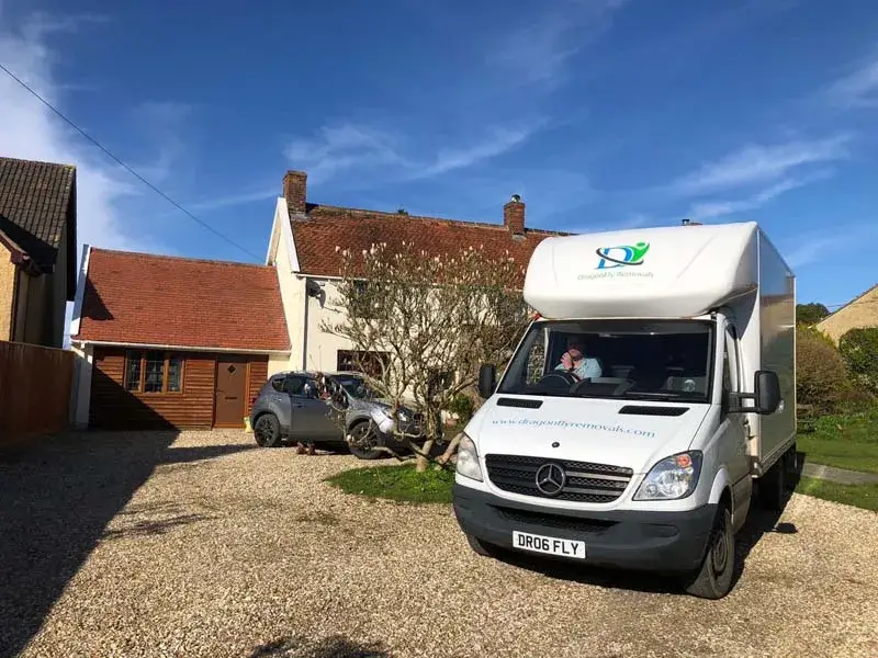 A white delivery truck is parked on a gravel driveway in front of a house with a brick roof. The truck has a logo on its side. A gray car is parked beside the house. The sky is clear and blue.