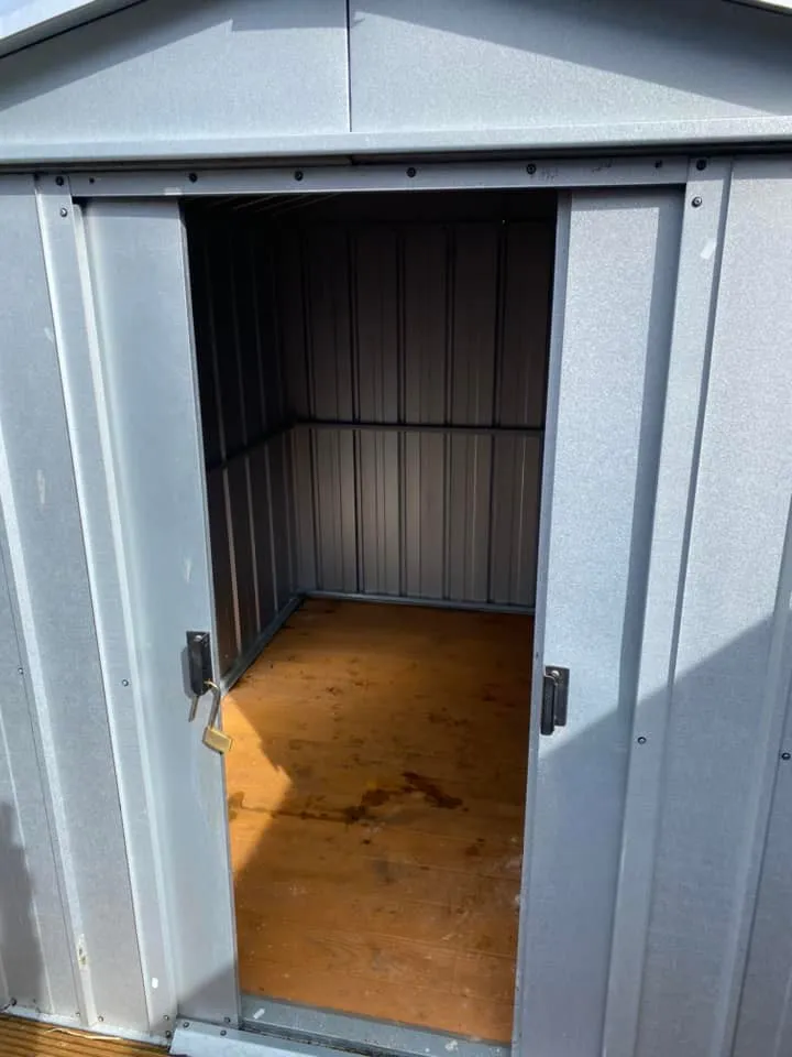 Open gray metal shed with a wooden floor, partially illuminated by sunlight. The shed is empty, showing clean and smooth interior walls. A padlock hangs on the handle of the open door.