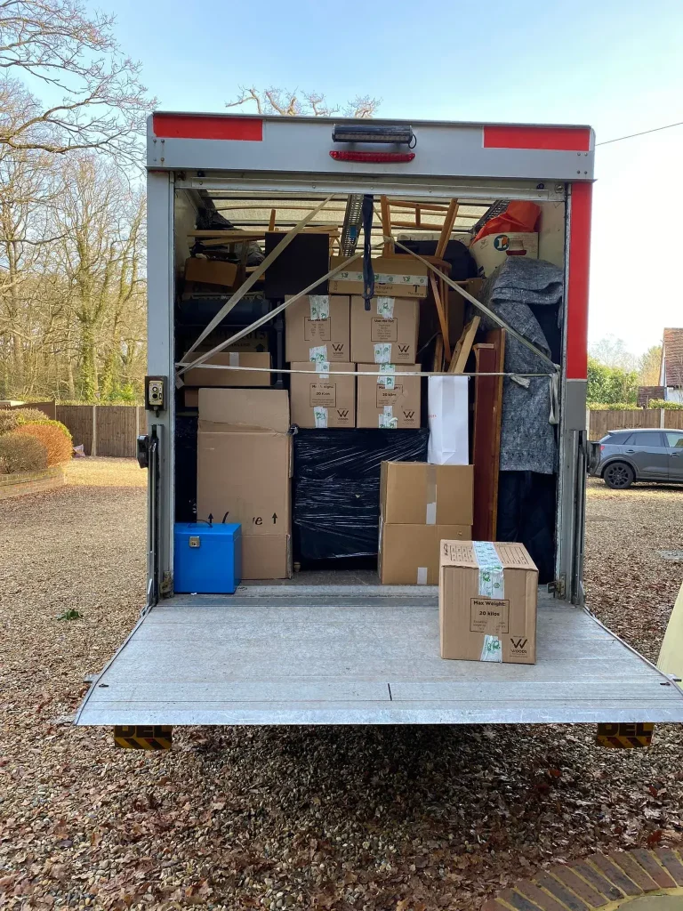 The image shows the open back of a moving truck filled with cardboard boxes and furniture, including chairs and wrapped items. A blue cooler and a labeled box are at the edge of the truck. The scene is outdoors with trees and a parked car nearby.