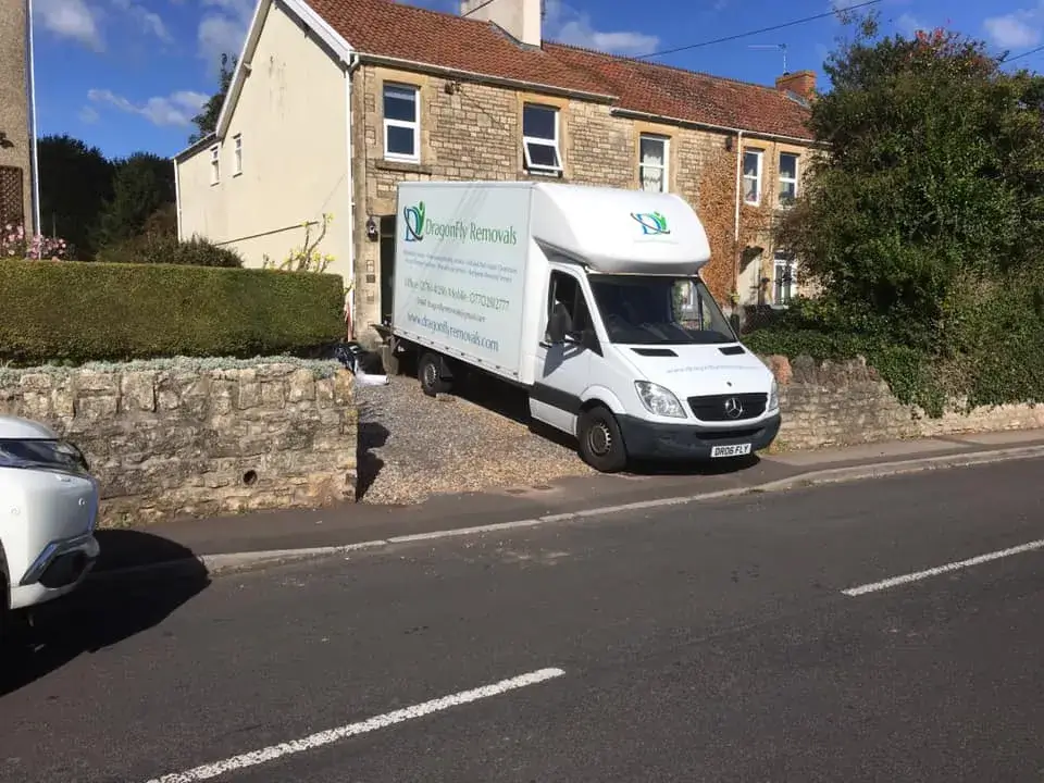 A white delivery van labeled "Dragonfly Networks" is stuck in a narrow gravel driveway between a stone house and a hedge, partially on the sidewalk, with its rear jutting onto a two-lane road. The sky is clear with a few clouds.