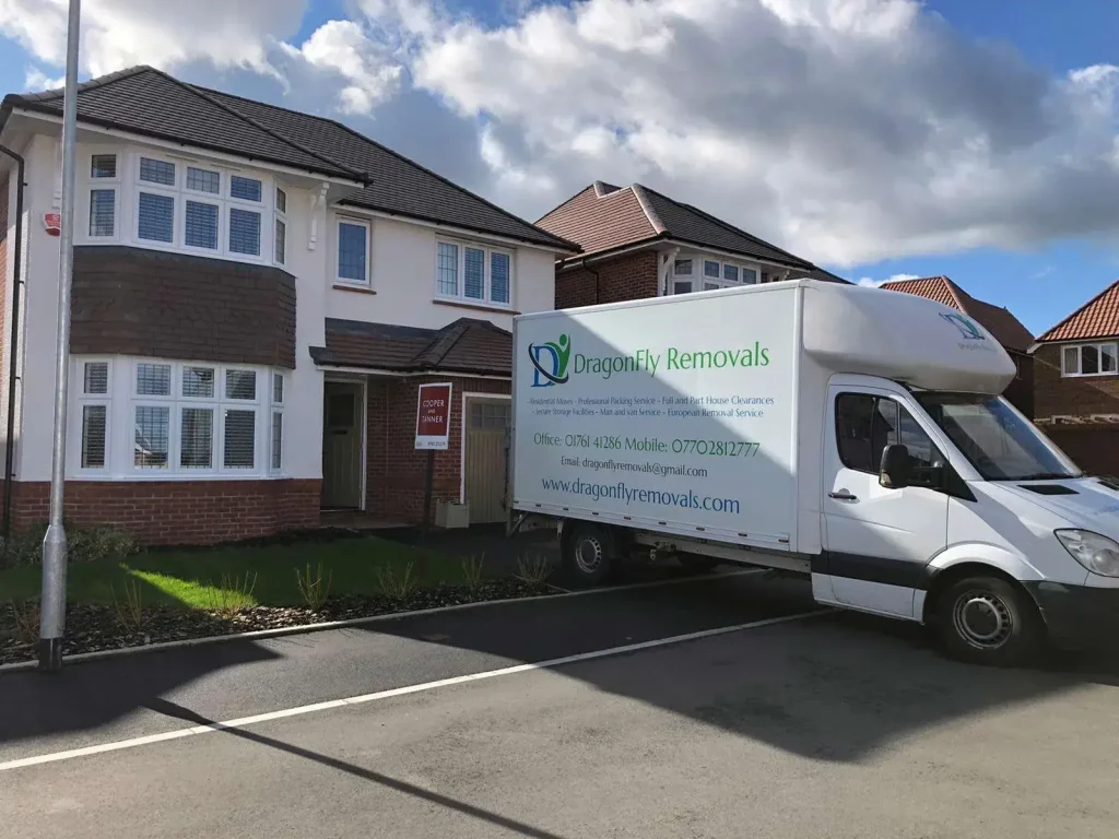 A white moving truck with "DragonFly Removals" branding is parked in front of a suburban house with a brick and white facade. The truck's side displays contact information. A blue sky with scattered clouds is above.