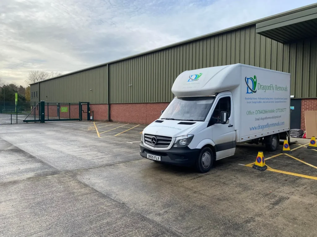 A white delivery truck labeled "Dragonfly Removals" is parked outside a large warehouse with green metal siding and red brickwork. The sky is cloudy, and there’s a fenced area with a gate to the left. Yellow road markings are visible on the ground.