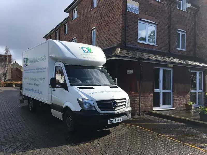 White delivery truck with "dignityfunerals.co.uk" and phone number branding, parked outside a three-story brick building on a cloudy day. The truck is a Mercedes-Benz, and the building has dark windows and a small sign above the entrance.