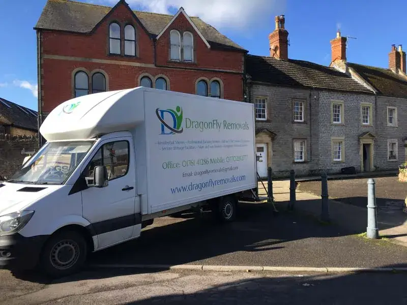 White moving truck with "DragonFly Removals" branding parked on a street with stone houses in the background. The sky is clear and blue, casting shadows on the road and buildings.