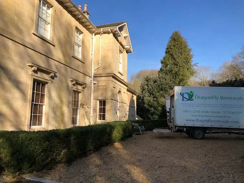 A moving truck with "Dragonfly Removals" parked in the driveway of a large stone house. The house is bordered by hedges, and there are trees in the background under a clear blue sky.