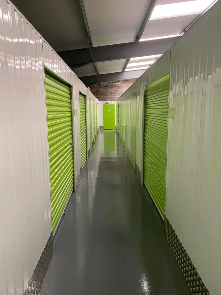 A long corridor in a storage facility with shiny gray floors and rows of units on either side. Units have bright green roll-up doors. The ceiling is metallic with overhead lighting, and the end of the corridor has a green door.