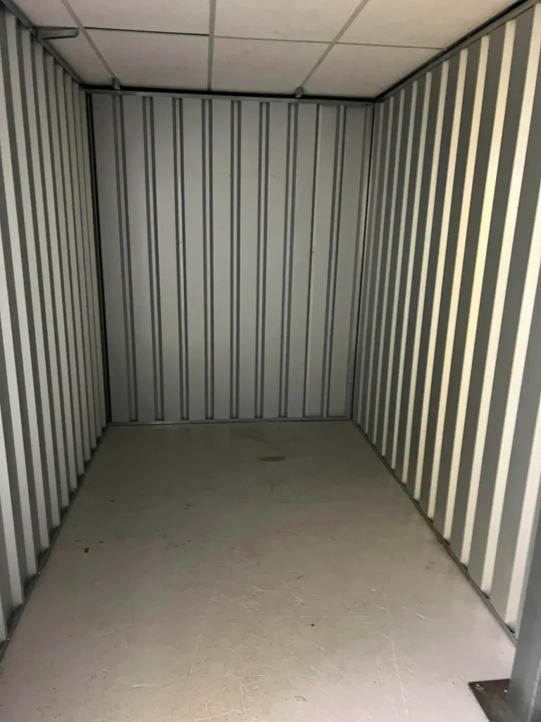 Empty storage unit with metal walls and ceiling. The floor is bare and appears to be concrete. The unit is well-lit from an overhead light, creating a clean and organized appearance.